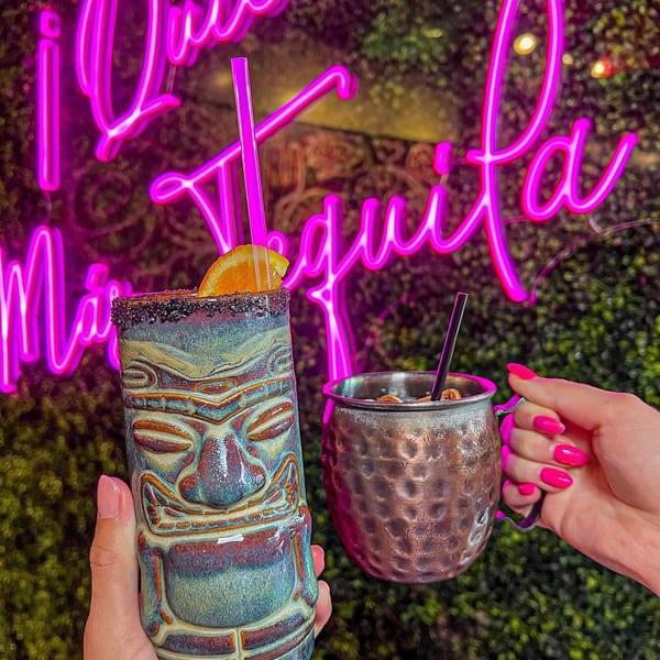 Two hands holding cocktails, a tiki mug and a copper mug, in front of a pink neon sign.