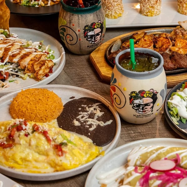 A table laden with various Mexican dishes, including fajitas, enchiladas, and drinks in Kahlolera mugs.