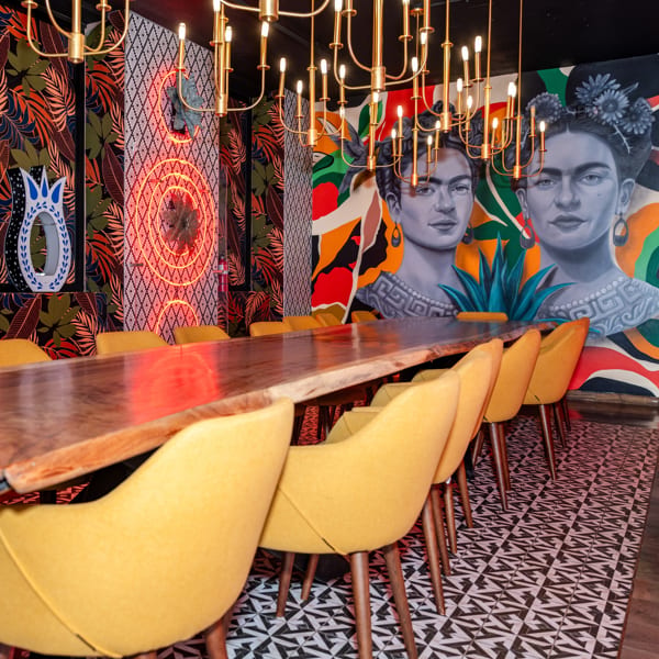 Vibrant restaurant interior with a long wooden table, yellow chairs, Frida Kahlo murals, and gold chandeliers.
