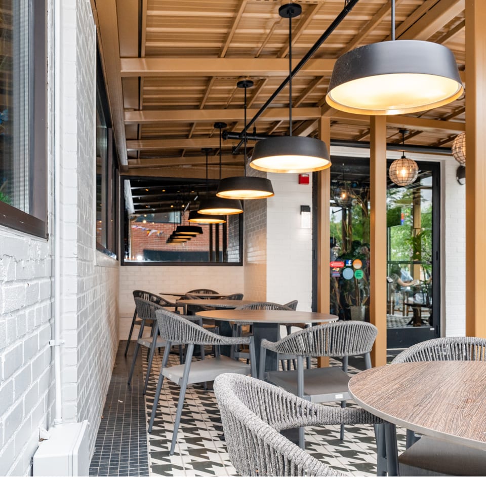 Modern outdoor restaurant patio with grey woven chairs, round tables, and black and white patterned floor.