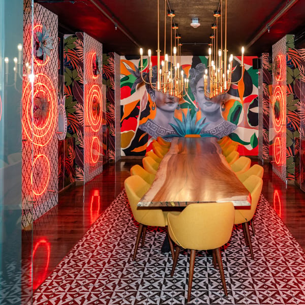 Colorful restaurant dining room featuring a long table, yellow chairs, a mural, and red neon lighting.
