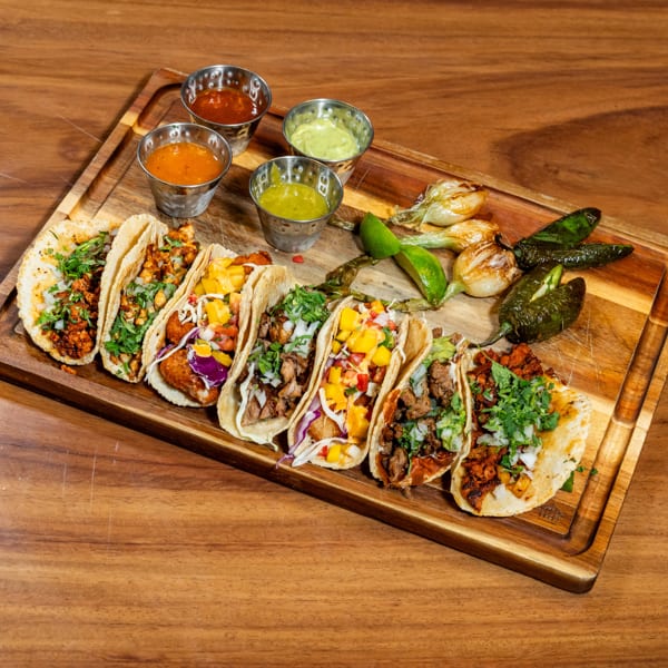Assortment of colorful tacos with various fillings, sauces, grilled onions, and peppers on a wooden board.