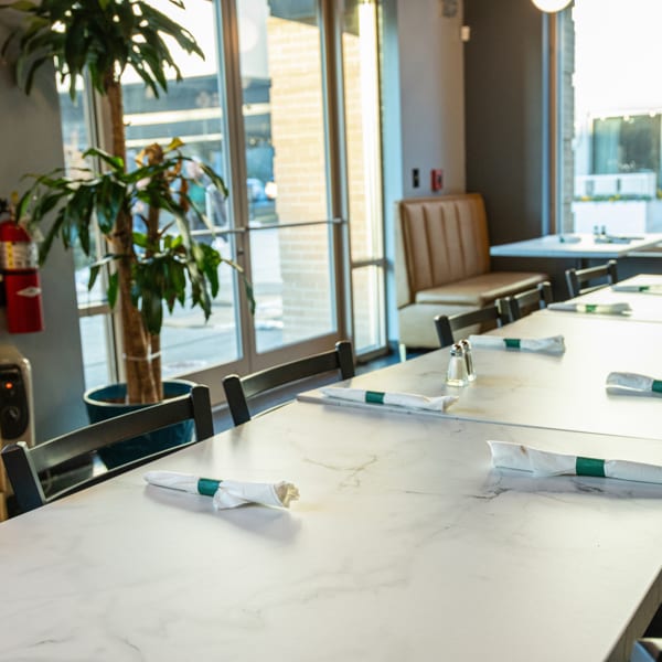 Bright restaurant interior featuring long white tables, chairs, booth seating, and large windows.