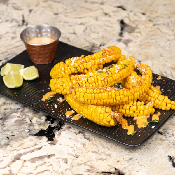 Elote-style corn on the cob with sauce, cheese, and lime wedges on a black platter.