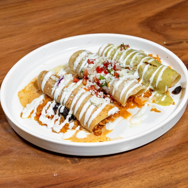 Three enchiladas with green and orange sauces, topped with crema, pico de gallo, and cheese on a white plate.