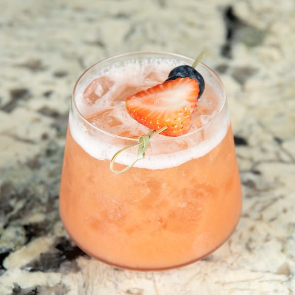 A refreshing pink cocktail with ice, garnished with a strawberry half and a blueberry on a skewer.