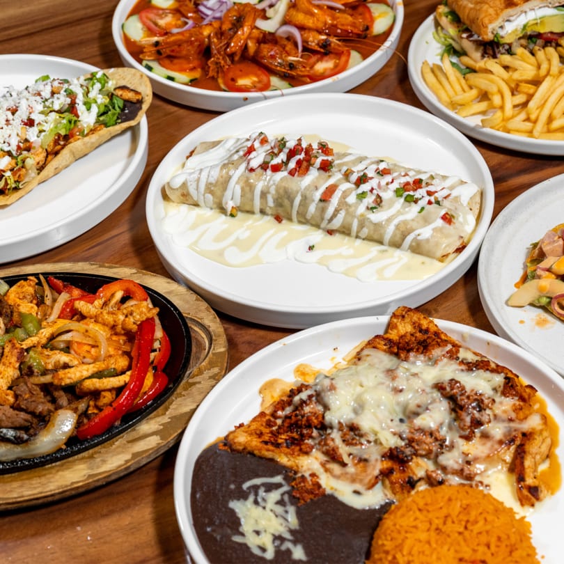 A wooden table laden with various Mexican dishes, including a burrito, fajitas, tacos, and shrimp.