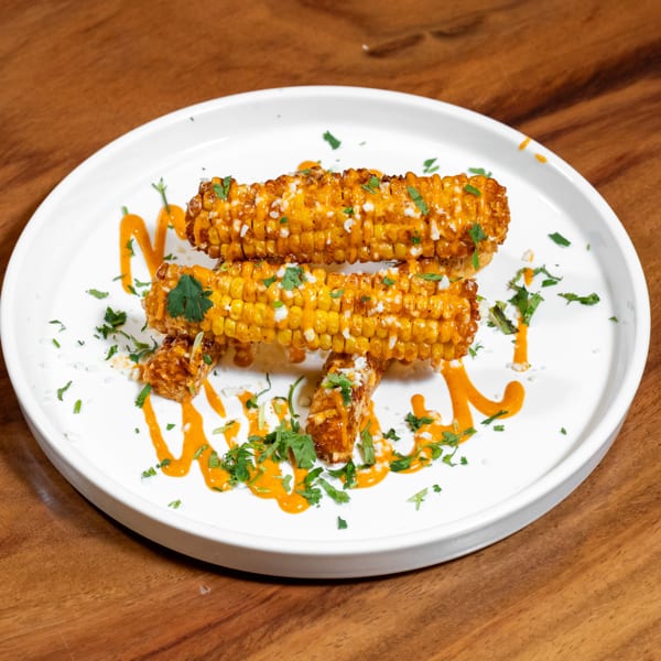 Roasted corn on the cob pieces, drizzled with orange sauce, crumbled cheese, and fresh cilantro on a white plate.