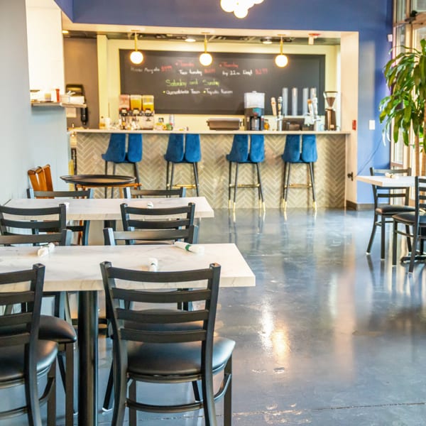 Empty restaurant dining room with white tables, black chairs, and a bar with blue stools and a chalkboard menu.