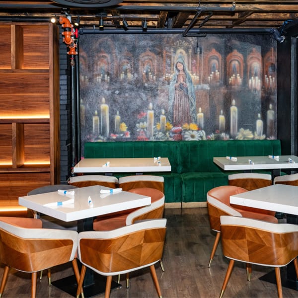 Modern restaurant interior with white tables, wood chairs, green velvet banquette, and a mural of a praying figure with candles.