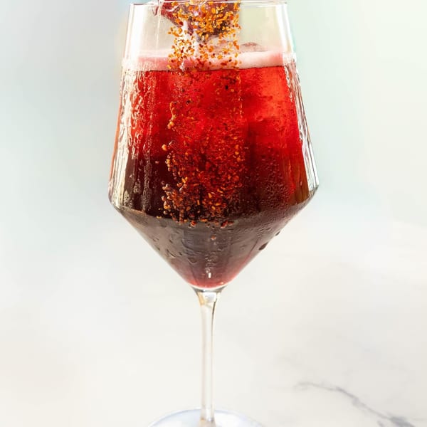 Red cocktail in a wine glass with a bottle pouring in golden sprinkles.