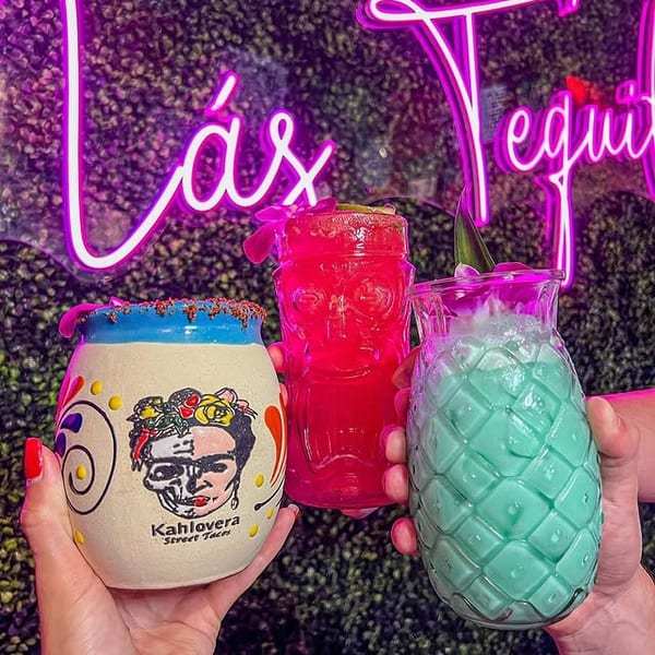 Three colorful cocktails, including a Frida Kahlo mug, held against a neon-lit green wall.