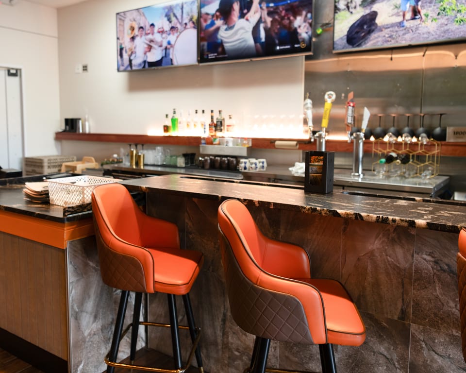 Empty bar area with orange bar stools, a dark marble counter, and TVs displaying sports.