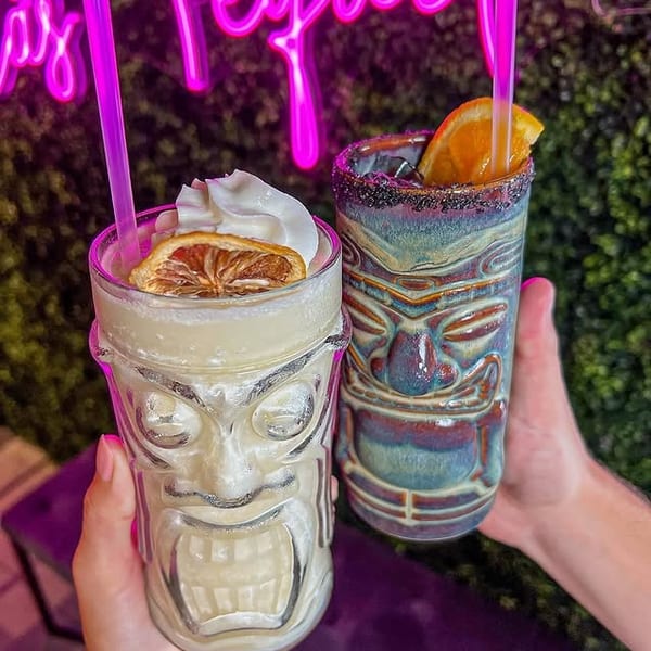 Two hands hold tiki drinks with garnishes, against a neon pink sign and green foliage.