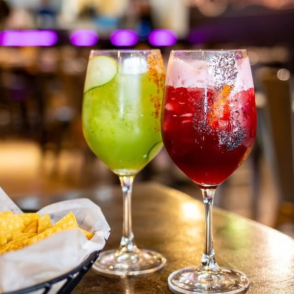 Two colorful cocktails, one green and one red, with a basket of tortilla chips on a table.