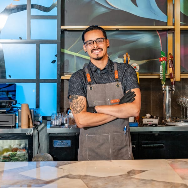 Smiling bartender with glasses and apron stands behind a bar with arms crossed.