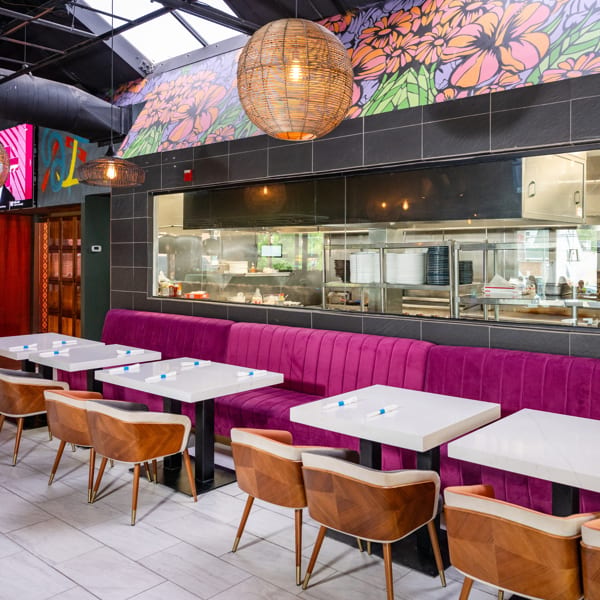 Restaurant interior with purple booth seating, white tables, wooden chairs, and a colorful floral mural.