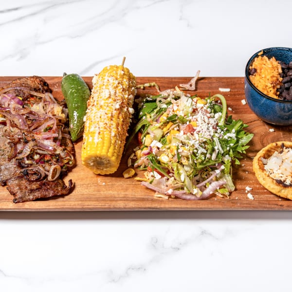 Grilled steak with onions, elote, salad, rice, black beans, and a corn cake on a wooden board.