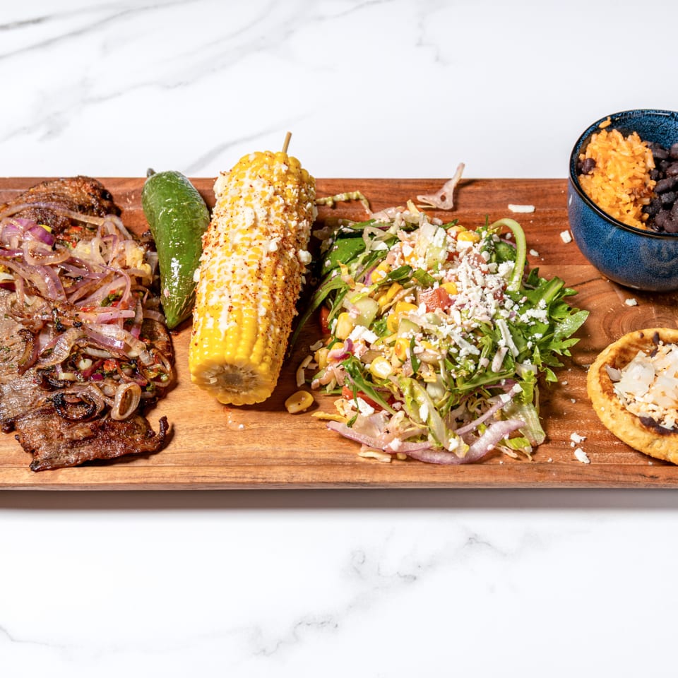 Mexican-inspired platter with grilled steak, street corn, salad, cucumber, tortilla, and bowls of rice and black beans.
