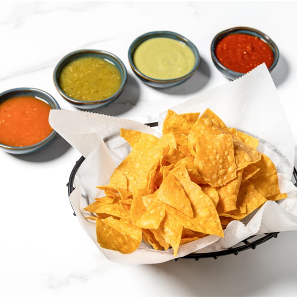 Crispy tortilla chips in a basket with four colorful salsa dips on a white marble surface.
