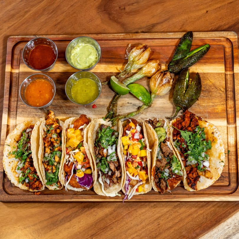 Assortment of tacos with various fillings, sauces, grilled onions, and peppers on a wooden board.