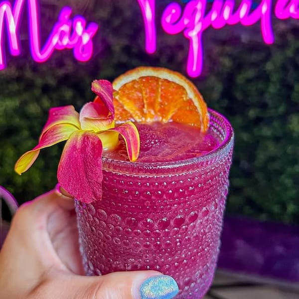 Hand holding a vibrant pink frozen drink garnished with an orange slice and orchid, under a neon sign.