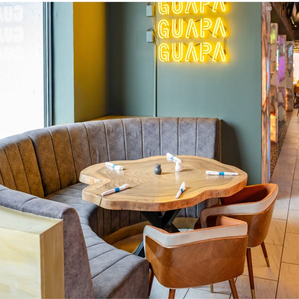 Modern restaurant interior with a live-edge wooden table, gray booth seating, and a neon 'GUAPA' sign.