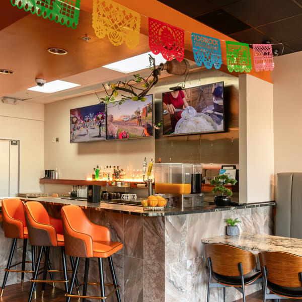 Restaurant bar area with orange bar stools, multiple TVs, and colorful papel picado decorations hanging above.