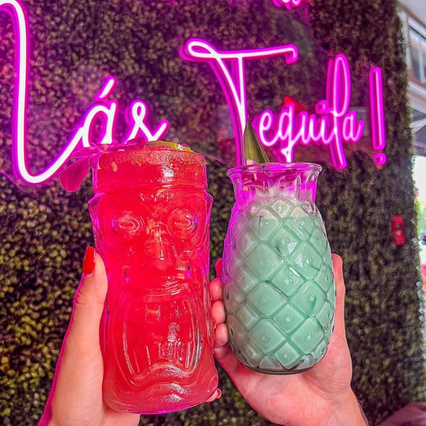 Two hands hold a red tiki drink and a green pineapple drink against a neon sign.