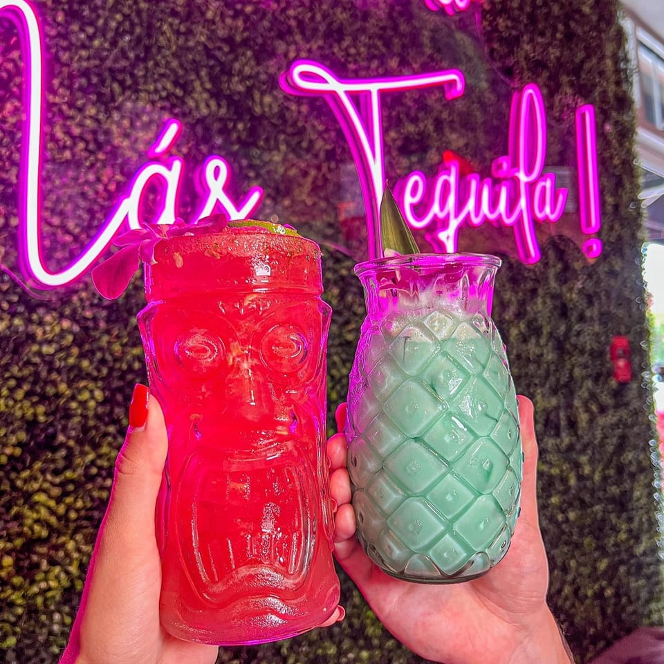 Two hands hold a red tiki drink and a green pineapple drink against a neon sign.