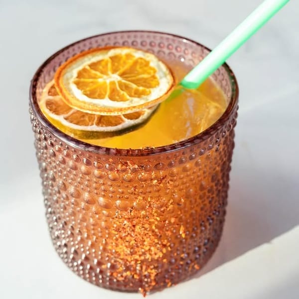 A vibrant orange cocktail in a textured glass, garnished with dried orange slices and a green straw.