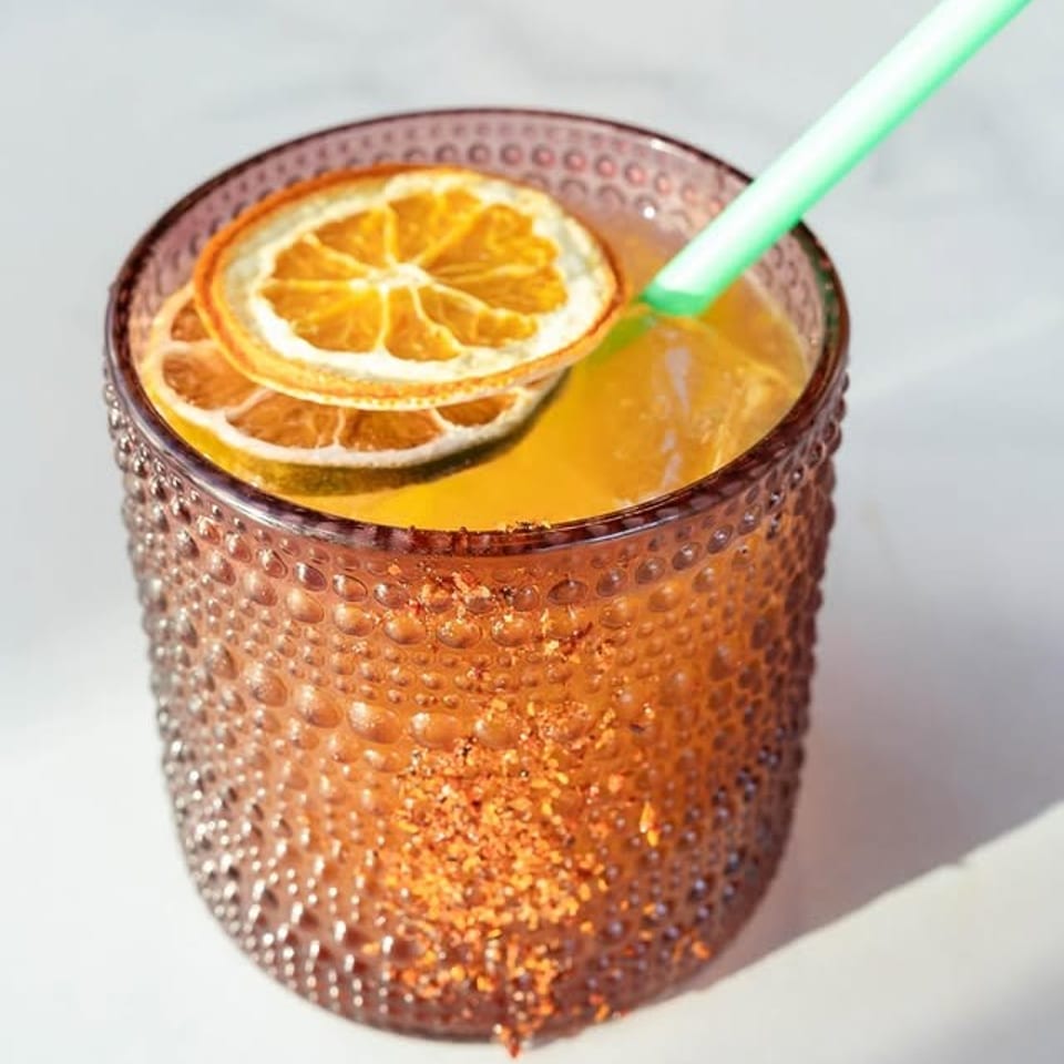 A vibrant orange cocktail in a textured glass, garnished with dried orange slices and a green straw.
