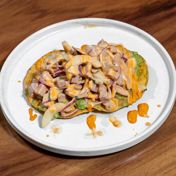 Tostada topped with ceviche, avocado, red onions, and creamy orange sauce on a white plate.