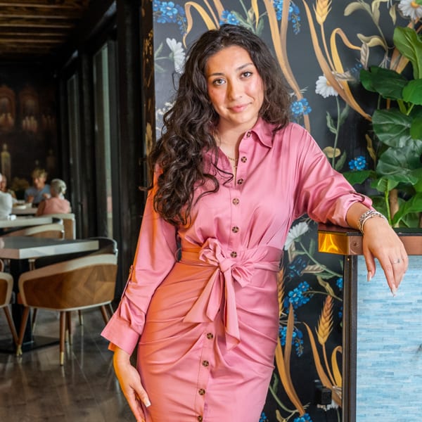 Woman in a pink dress smiling, leaning on a counter in a restaurant with floral wallpaper.