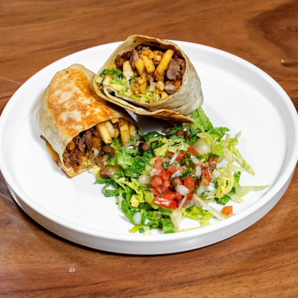 Two halves of a burrito filled with meat and fries, served with a fresh side salad on a white plate.