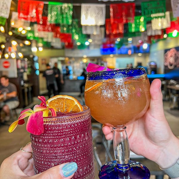 Two hands holding cocktails with orange slices and orchids, under colorful papel picado decorations.