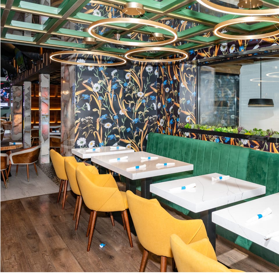 Modern restaurant interior with yellow chairs, green booth, floral wallpaper, and gold circular ceiling lights.